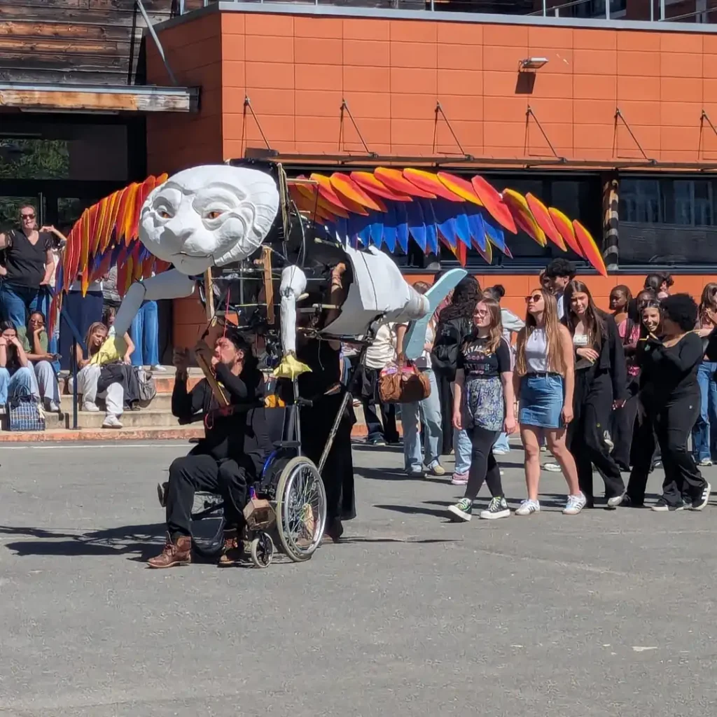 Dans la cour du lycée Jean‑Zay, une grande marionnette articulée représentant une créature fantastique à tête blanche et ailes colorées déambule. La marionnette est manipulée par plusieurs personnes, dont l’une est assise dans un fauteuil roulant et actionne des commandes reliées à la structure. Autour de la marionnette, un groupe d’élèves marche et observe la scène. La déambulation se déroule en plein jour, devant une façade orange du lycée, dans un cadre collectif et festif.