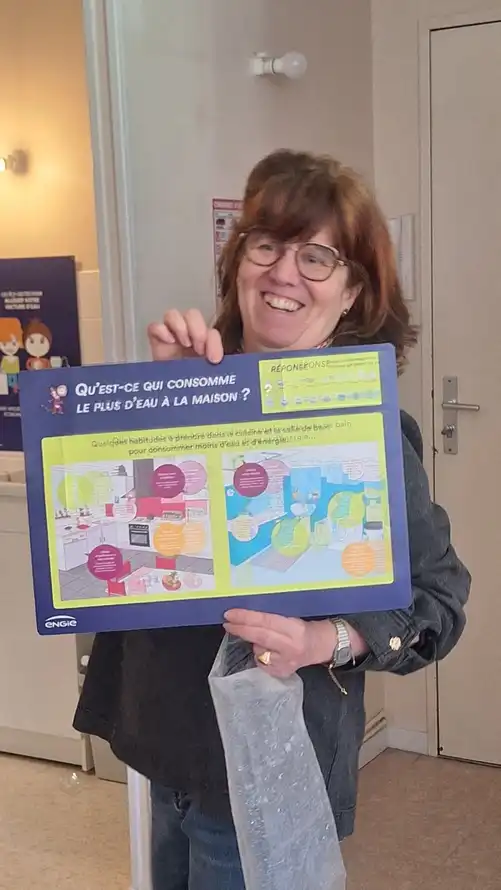 Une personne se tenant dans un appartement pédagogique présente un panneau d’information intitulé « Qu’est-ce qui consomme le plus d’eau à la maison ? ». Le support illustré compare les usages domestiques, notamment la cuisine, la salle de bain et les toilettes, et explique les postes les plus consommateurs d’eau. L’affiche met en avant des conseils pratiques pour réduire la consommation d’eau au quotidien.